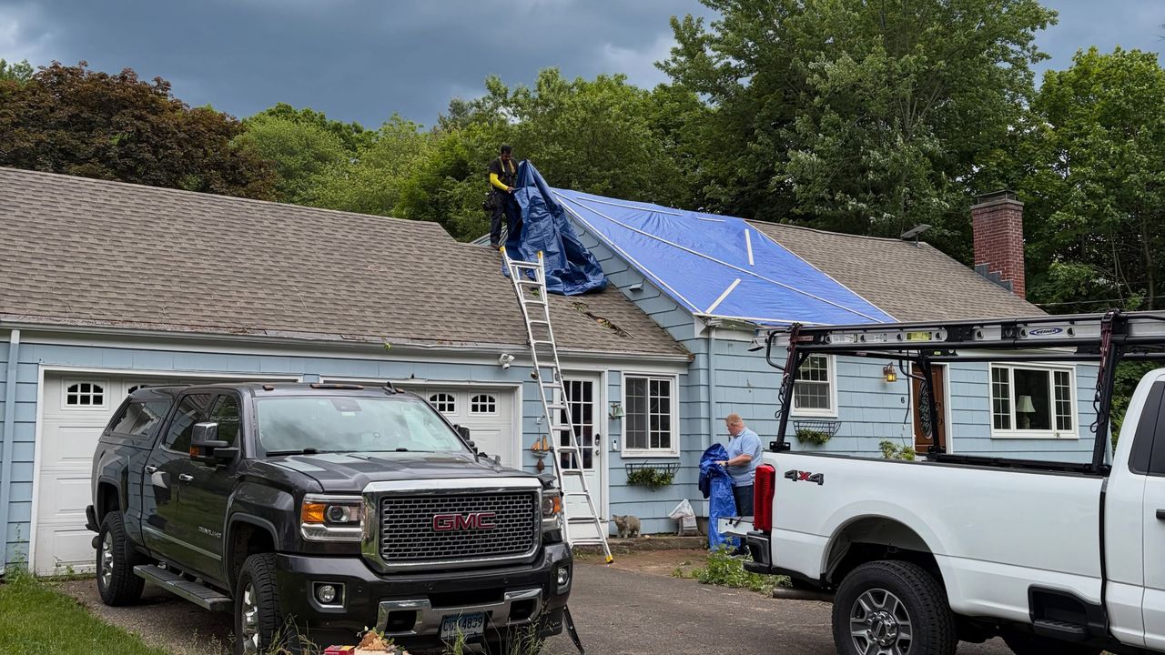 The same house after the tree came off: someone on a ladder spreading a blue tarp over the damaged roof, trucks in the drive.